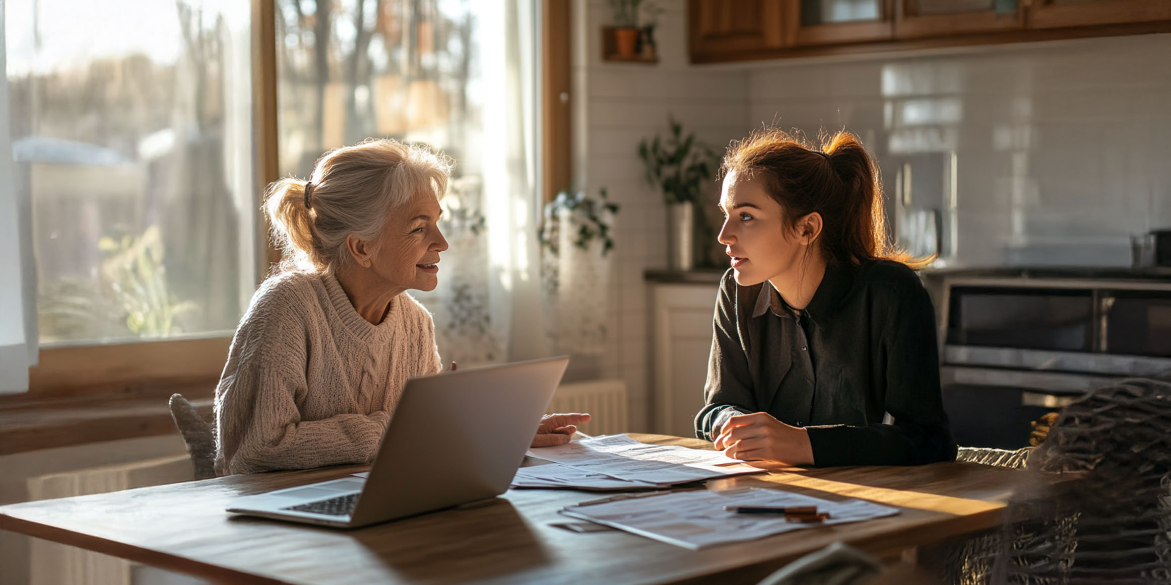 Senior woman talking with female Medicare insurance agent at kitchen table with laptop and paperwork